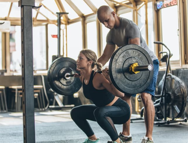 A trainer helps a woman lift heavy weights